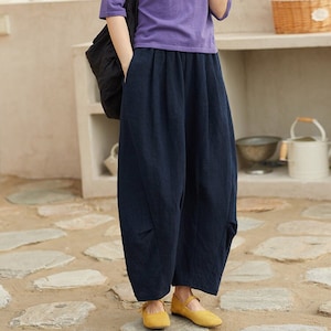 May include: A woman wearing a purple short-sleeved shirt with a black and white polka dot scarf, dark blue wide-leg pants, and yellow flats.