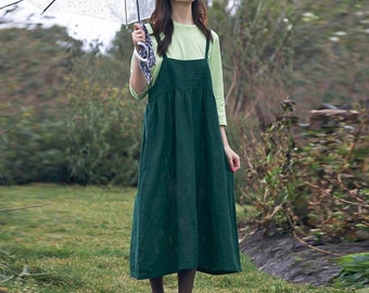 May include: A woman wearing a green linen overall dress with thin straps. The dress has a gathered waist and a midi length. She is holding a clear umbrella and standing in a grassy area.