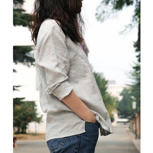 May include: A light grey linen shirt with long sleeves and a button-up front. The shirt is being worn by a person with long dark hair and is tucked into blue jeans.