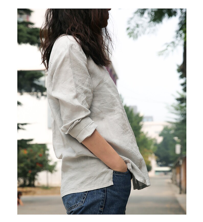 May include: A light grey linen shirt with long sleeves and a button-up front. The shirt is being worn by a person with long dark hair and is tucked into blue jeans.