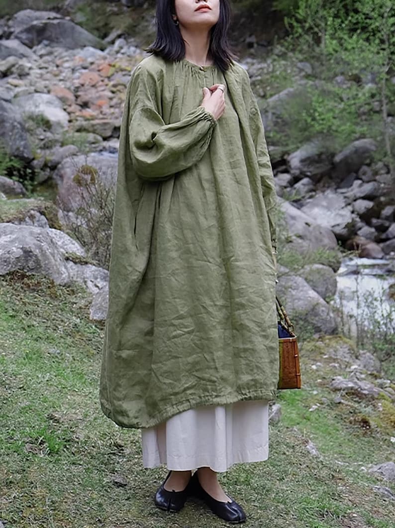 May include: A loose-fitting, long-sleeved, green linen dress with a gathered neckline and a slightly gathered skirt. The dress is worn by a person standing in a natural setting.