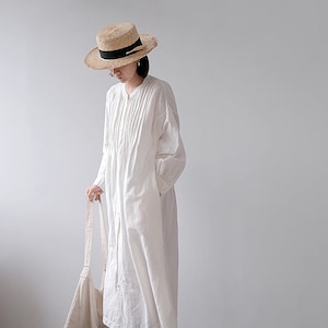 May include: A white long-sleeved shirt dress with a button-up front and a straw hat with a black ribbon. The person is holding a white tote bag.