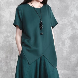 May include: A dark green, loose-fitting, short-sleeved top with a slightly asymmetrical hemline. The top has a round neckline and features a long, black beaded necklace.