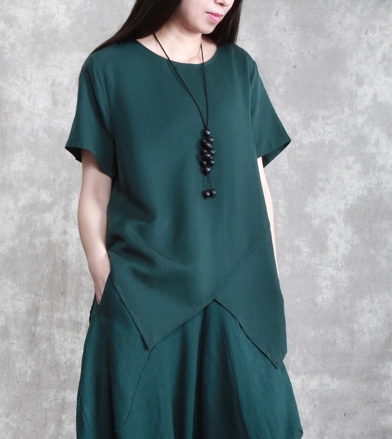 May include: A dark green, loose-fitting, short-sleeved top with a slightly asymmetrical hemline. The top has a round neckline and features a long, black beaded necklace.