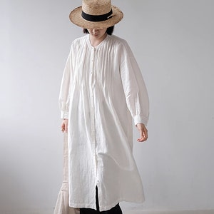 May include: A white long-sleeved shirt dress with a button-up front and a pleated bodice. The dress is worn with black trousers and black shoes. The person is wearing a straw hat with a black ribbon.