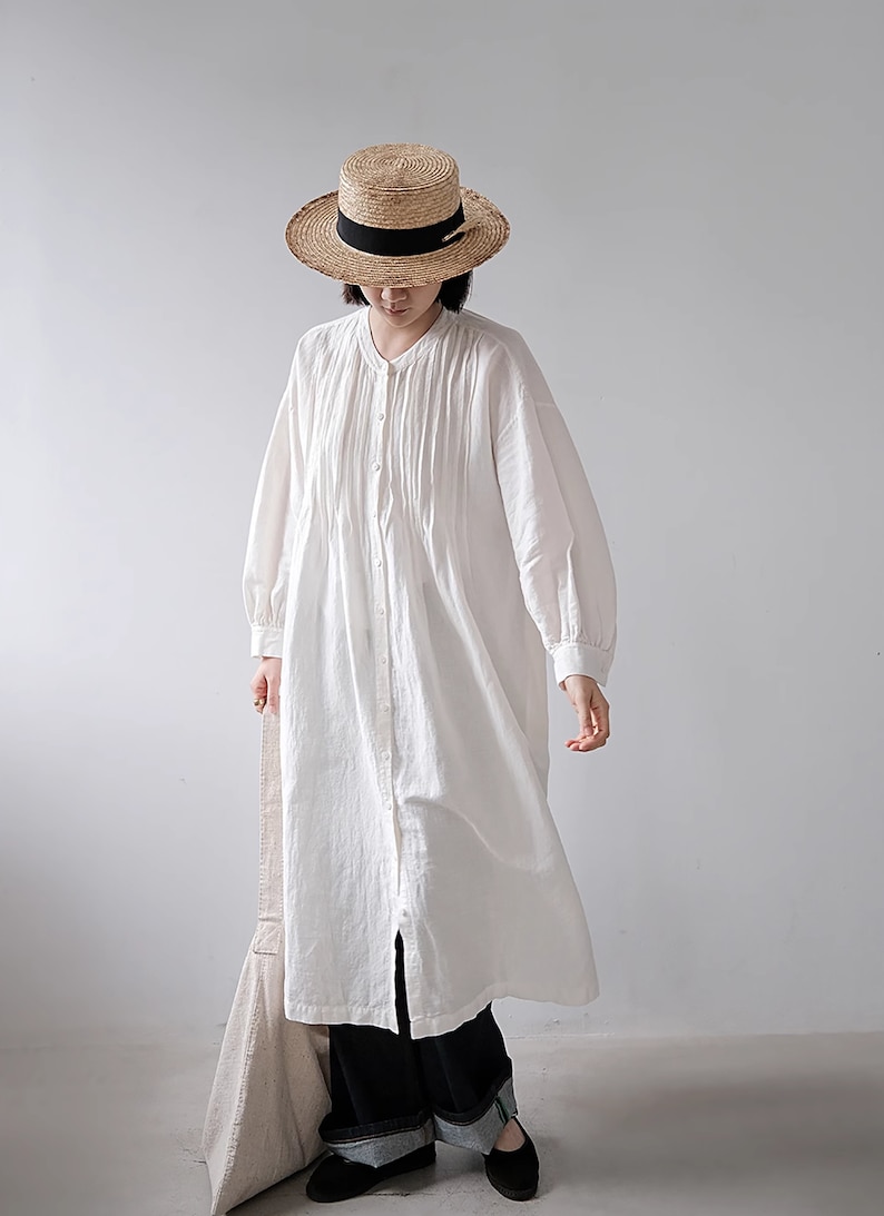 May include: A white long-sleeved shirt dress with a button-up front and a pleated bodice. The dress is worn with black trousers and black shoes. The person is wearing a straw hat with a black ribbon.