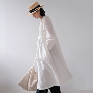May include: A white linen dress with long sleeves and a gathered waist. The dress is paired with dark blue jeans and brown shoes. The person is wearing a straw hat with a black ribbon.