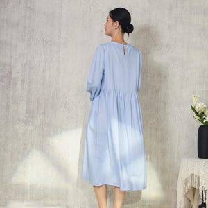 May include: A light blue dress with long sleeves and a gathered waist. The dress has a keyhole detail at the back of the neck. The person is wearing brown sandals. The background is a textured, light gray wall.