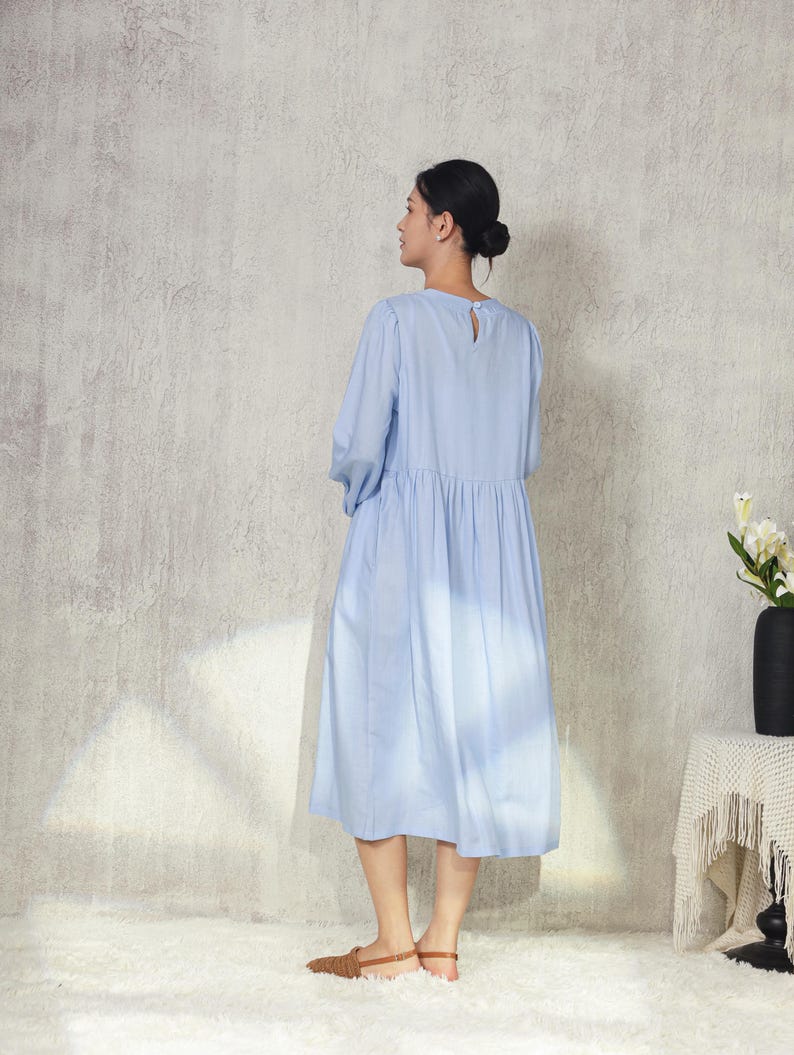 May include: A light blue dress with long sleeves and a gathered waist. The dress has a keyhole detail at the back of the neck. The person is wearing brown sandals. The background is a textured, light gray wall.
