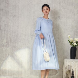 May include: A light blue long-sleeve dress with a round neckline and a gathered skirt. The dress is paired with a small white purse with a long strap. The person is wearing brown sandals. A black vase with white flowers sits on a table.