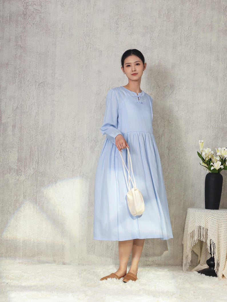 May include: A light blue long-sleeve dress with a round neckline and a gathered skirt. The dress is paired with a small white purse with a long strap. The person is wearing brown sandals. A black vase with white flowers sits on a table.