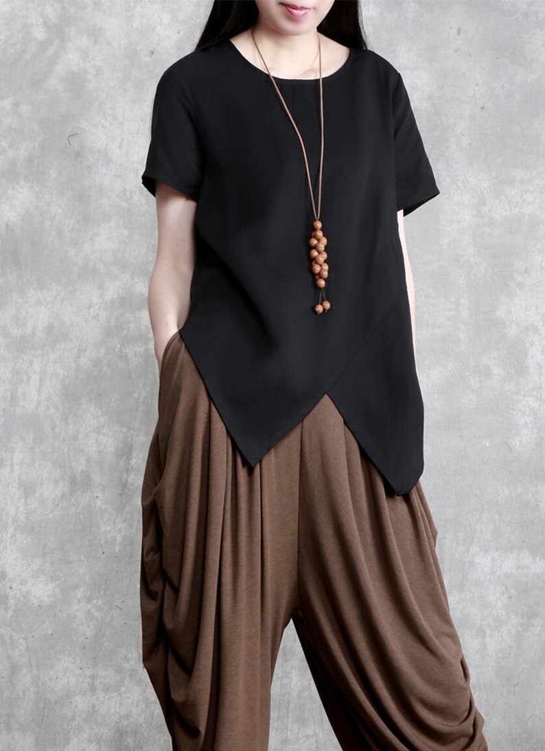 May include: A black, short-sleeved, loose-fitting top with a diagonal hemline. The top is paired with brown, loose-fitting pants. The model is wearing a long, brown beaded necklace.
