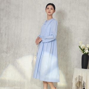 May include: A light blue long-sleeve dress with a round neckline and pleated details. The dress has a flowing skirt and is paired with woven sandals. A white purse hangs from the shoulder. A black vase with white flowers sits on a side table.