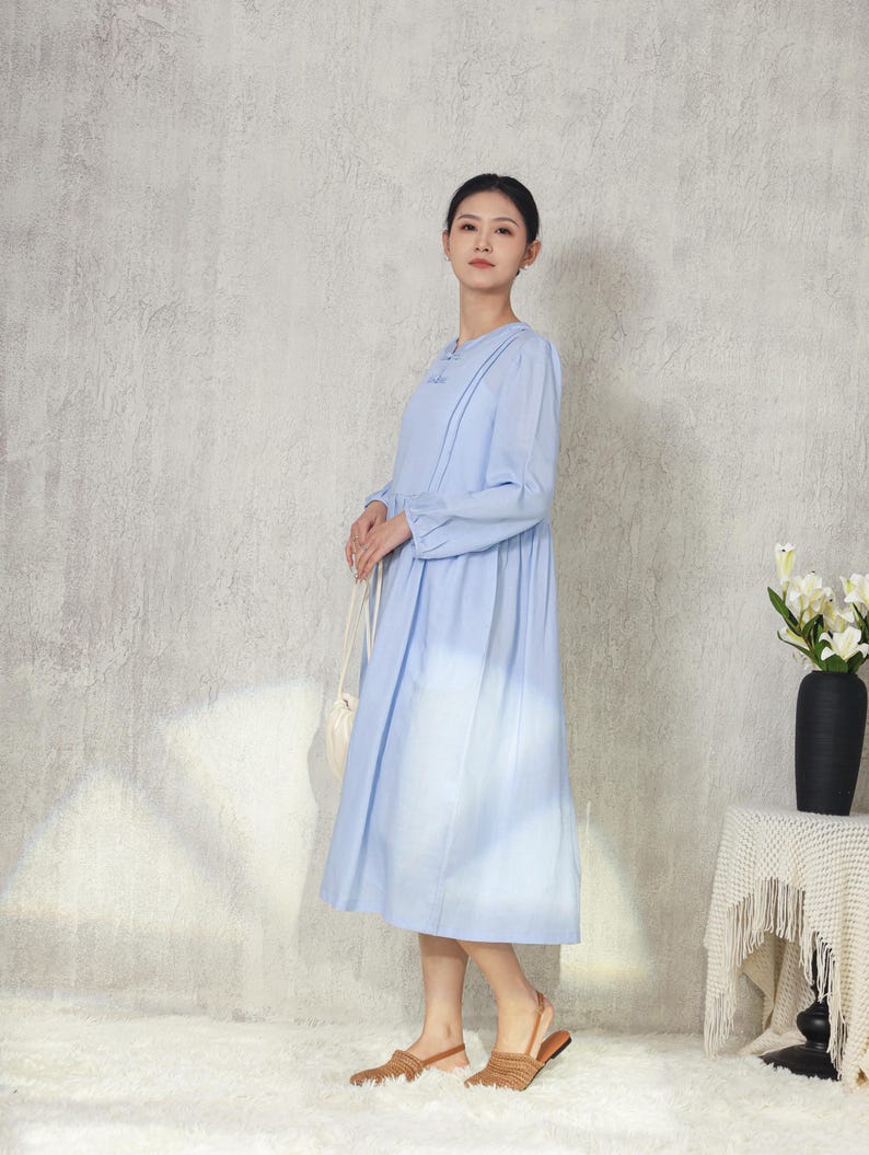 May include: A light blue long-sleeve dress with a round neckline and pleated details. The dress has a flowing skirt and is paired with woven sandals. A white purse hangs from the shoulder. A black vase with white flowers sits on a side table.