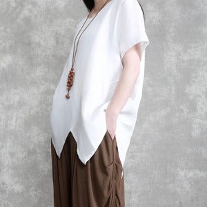 May include: A white linen top with short sleeves and a slightly asymmetrical hem. The top has a brown wooden bead necklace.