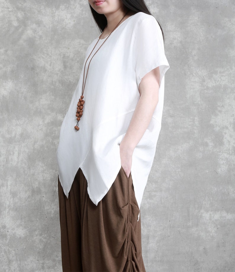 May include: A white linen top with short sleeves and a slightly asymmetrical hem. The top has a brown wooden bead necklace.