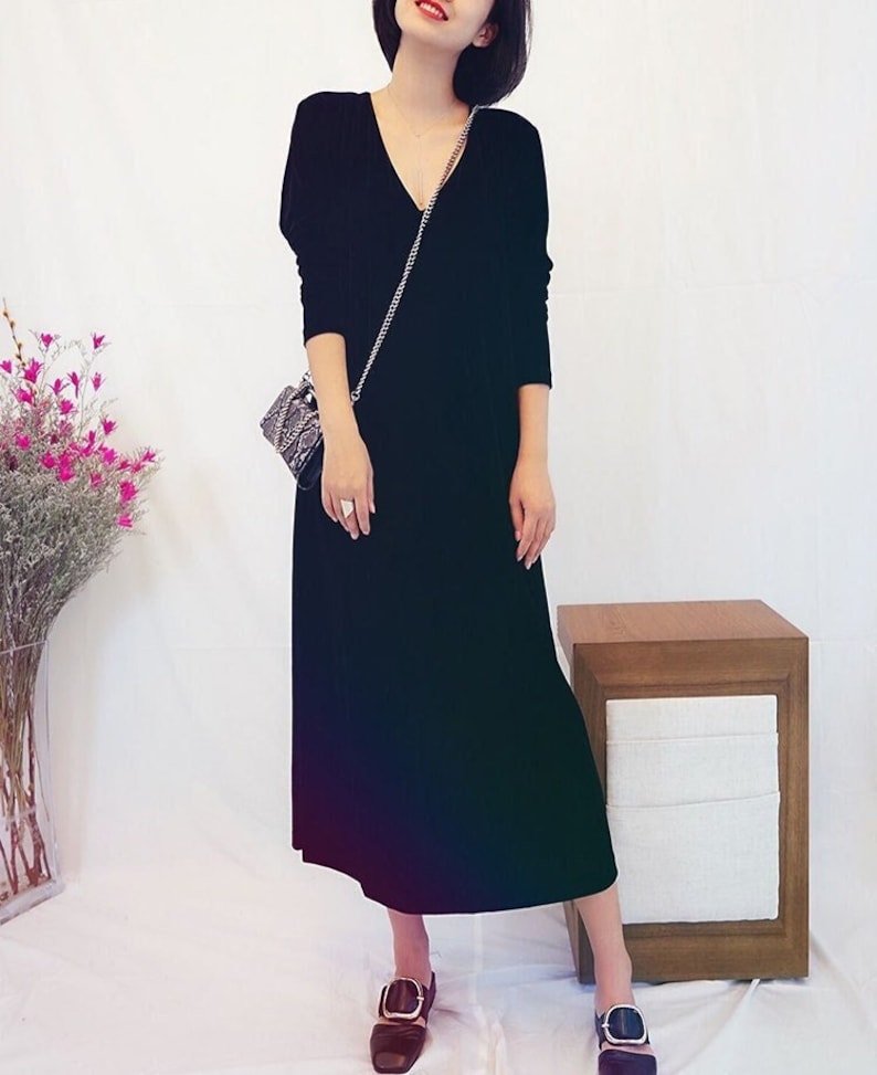 May include: A black long-sleeved midi dress with a v-neckline. The dress is made of a ribbed fabric and has a relaxed fit. The model is wearing the dress with a small black and silver chain bag and black shoes with gold buckles.