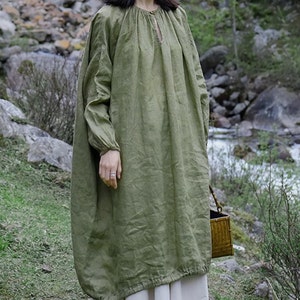 May include: A loose-fitting, long-sleeved, green linen dress with a gathered neckline and a keyhole detail. The dress is worn with a white skirt and black shoes.