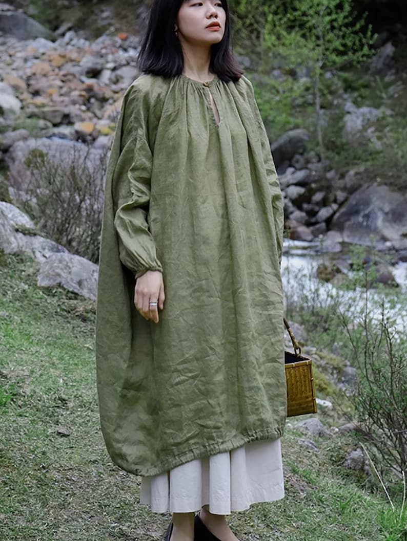 May include: A loose-fitting, long-sleeved, green linen dress with a gathered neckline and a keyhole detail. The dress is worn with a white skirt and black shoes.