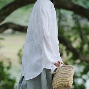 May include: A white linen shirt with long sleeves and a relaxed fit. The shirt is worn with a light green skirt and a woven straw bag.