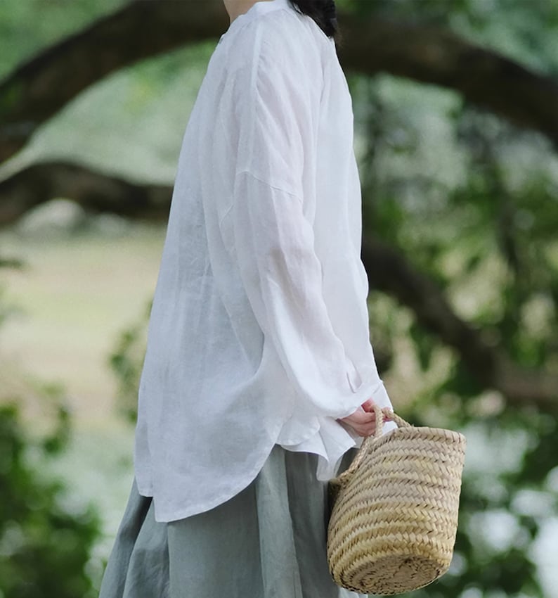 May include: A white linen shirt with long sleeves and a relaxed fit. The shirt is worn with a light green skirt and a woven straw bag.
