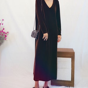 May include: A long brown velvet dress with a V-neckline and long sleeves. The dress is worn by a person with short dark hair and is paired with black shoes.