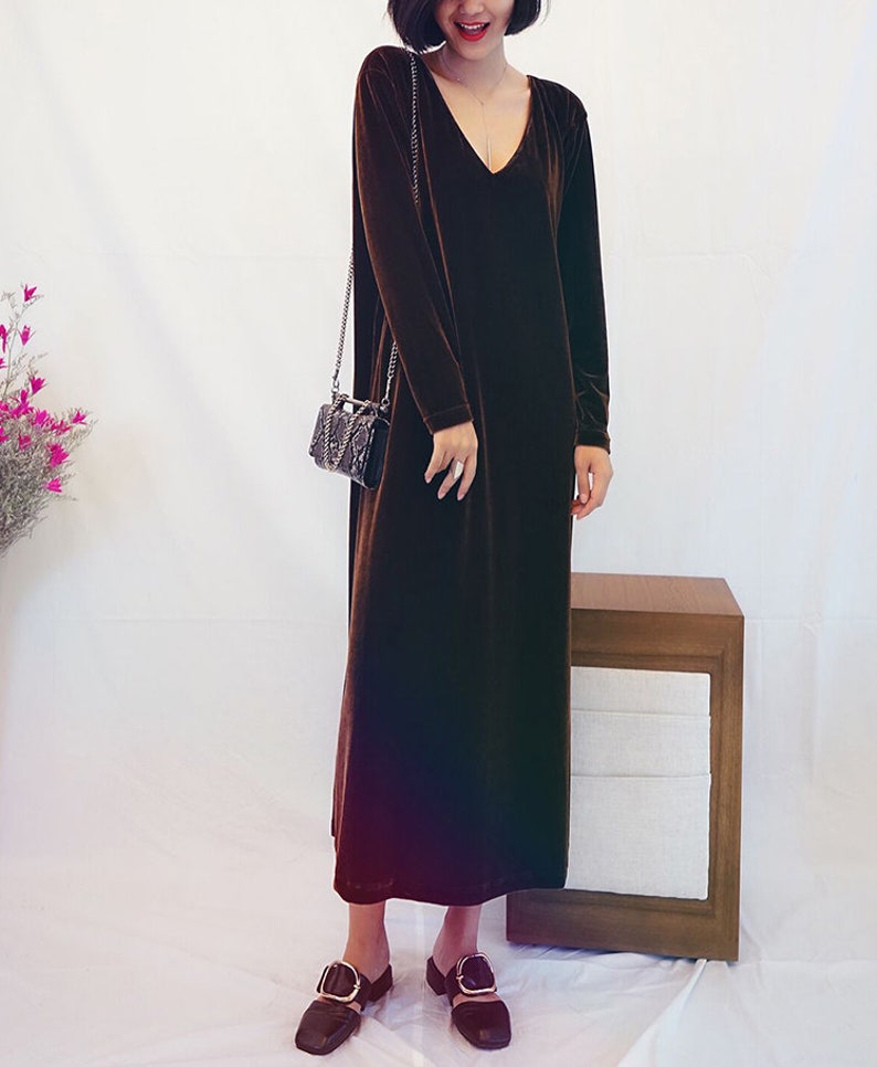 May include: A long brown velvet dress with a V-neckline and long sleeves. The dress is worn by a person with short dark hair and is paired with black shoes.