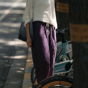 May include: A woman wearing a white shirt and purple linen pants. She is carrying a blue and white tote bag and standing next to a blue bicycle.