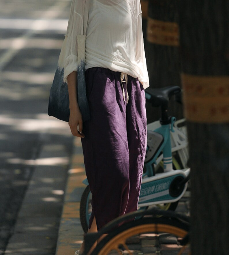 May include: A woman wearing a white shirt and purple linen pants. She is carrying a blue and white tote bag and standing next to a blue bicycle.