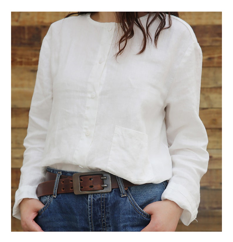May include: A white linen shirt with a button-up front and a single pocket on the left side. The shirt is worn with a brown leather belt with a silver buckle.