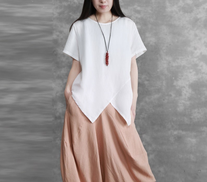 May include: A white linen top with a short sleeve and a diagonal hemline. The top features a red beaded necklace. The model is wearing a light brown skirt.