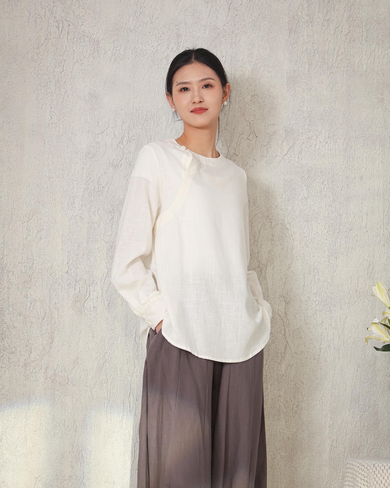 May include: Off-white long-sleeve blouse with a round neckline and a unique asymmetrical design. The blouse is paired with wide-leg, gray pants. The outfit is simple and elegant, suitable for casual or semi-formal wear.