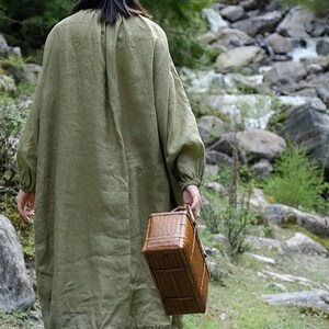 May include: A person wearing a green linen dress walks through a forest carrying a woven basket.