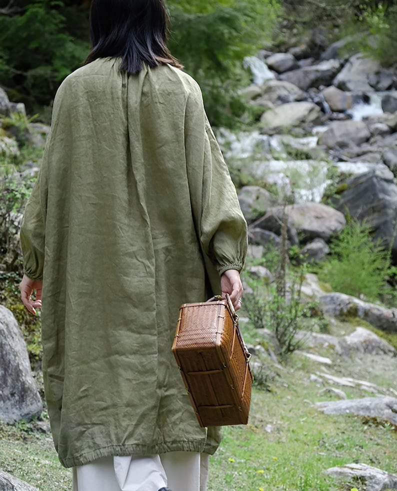 May include: A person wearing a green linen dress walks through a forest carrying a woven basket.