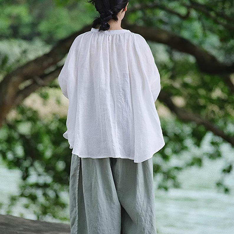 May include: A white, loose-fitting, long-sleeved blouse with a gathered neckline. The blouse is made of a lightweight fabric and has a relaxed fit. The model is wearing the blouse with a pair of light green pants.