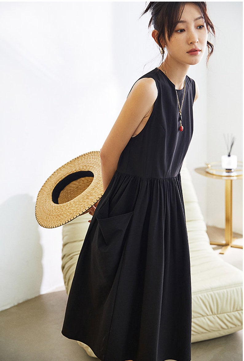 May include: A black sleeveless midi dress with a gathered waist and a large patch pocket on the left side. The dress is made of a lightweight fabric and has a relaxed fit.