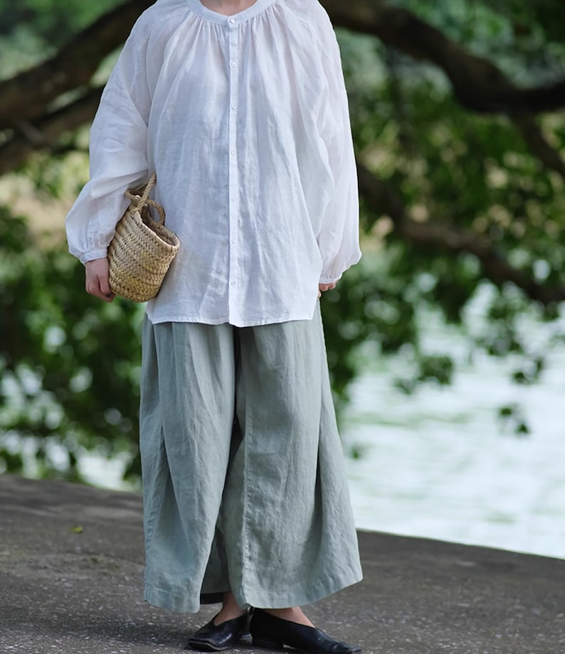 May include: A white linen blouse with a round neckline and long sleeves, paired with light green linen pants. The person is holding a small woven basket.