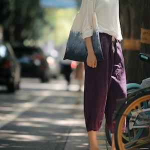 May include: A woman wearing a white shirt and purple pants is carrying a blue and white tie-dye tote bag. She is standing on a sidewalk with a bicycle in the background.