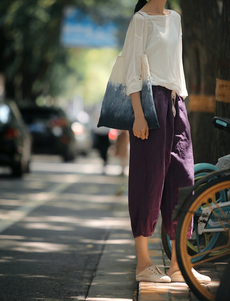 May include: A woman wearing a white shirt and purple pants is carrying a blue and white tie-dye tote bag. She is standing on a sidewalk with a bicycle in the background.