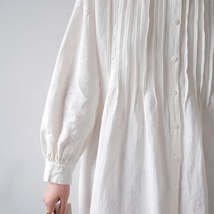 May include: A white linen shirt dress with a pleated front and long sleeves. The dress has a button-up closure and a relaxed fit.