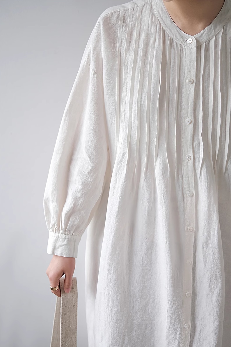 May include: A white linen shirt dress with a pleated front and long sleeves. The dress has a button-up closure and a relaxed fit.