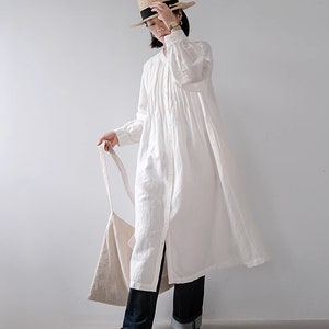 May include: A white linen dress with a button-down front and long sleeves. The dress has a gathered skirt and is worn with blue jeans and a straw hat.