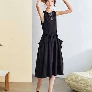 May include: A black sleeveless midi dress with two large pockets on the front. The dress has a gathered waist and a relaxed fit. The model is wearing the dress with black sandals and a straw hat.