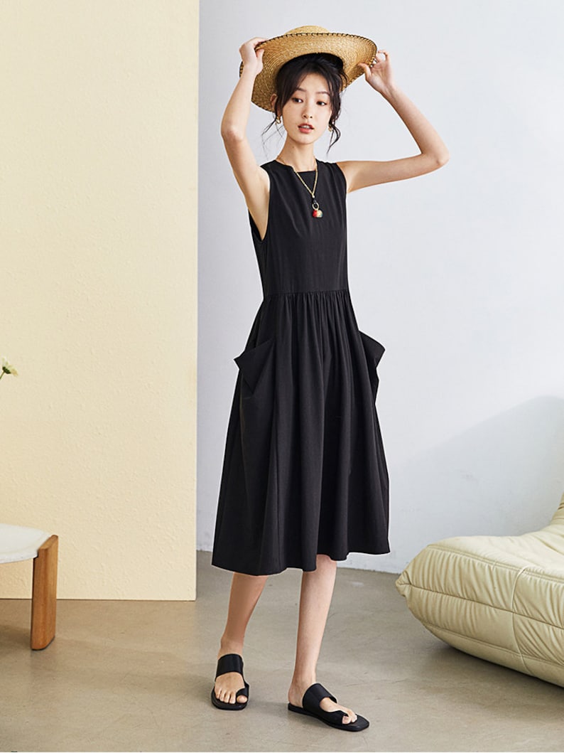May include: A black sleeveless midi dress with two large pockets on the front. The dress has a gathered waist and a relaxed fit. The model is wearing the dress with black sandals and a straw hat.