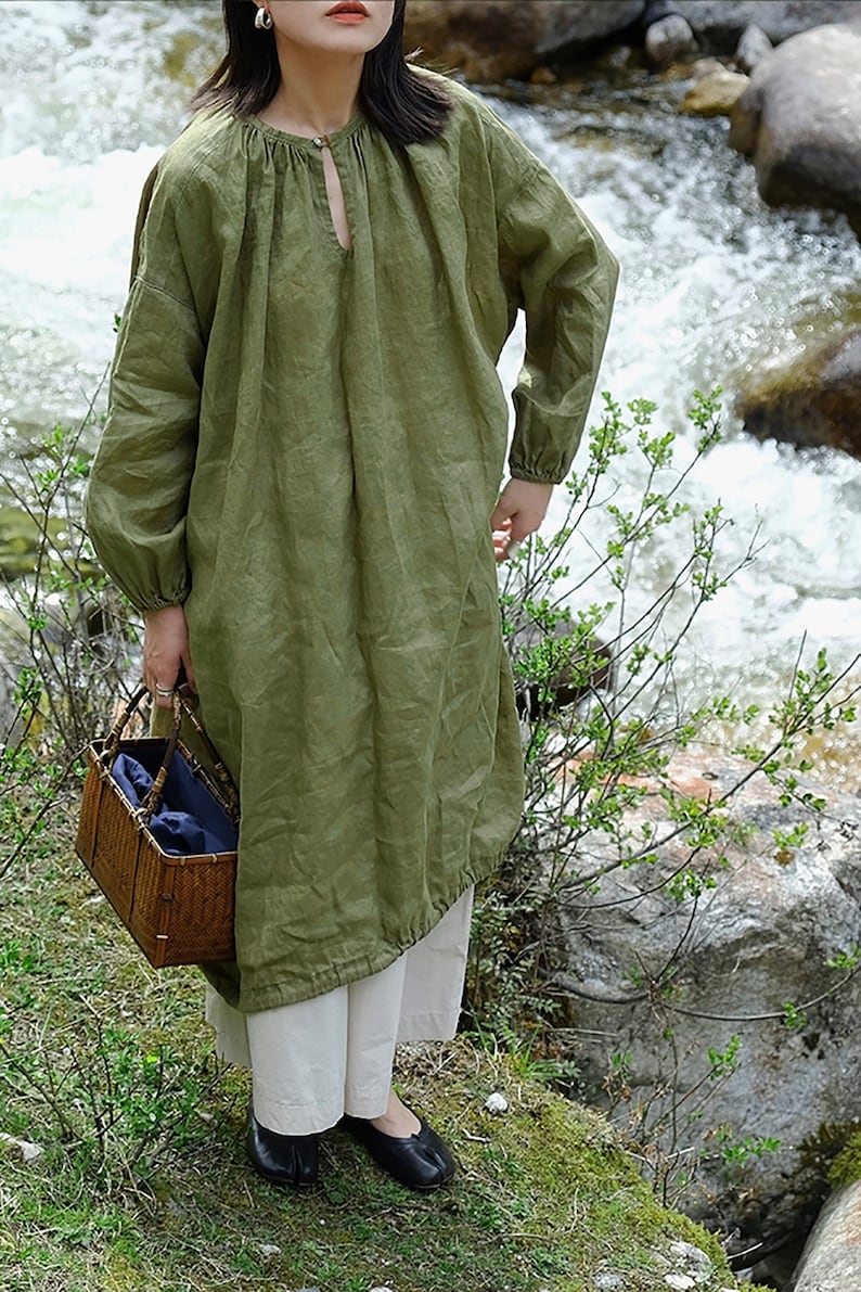 May include: A long-sleeved, green linen dress with a gathered neckline and a keyhole opening. The dress has a relaxed fit and is shown being worn by a person standing in a natural setting.