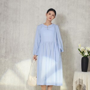 May include: A light blue dress with long sleeves and a round neckline. The dress has a gathered waist and falls to mid-calf. A small, off-white purse is held in the hand. The background is a light gray wall and a white rug.