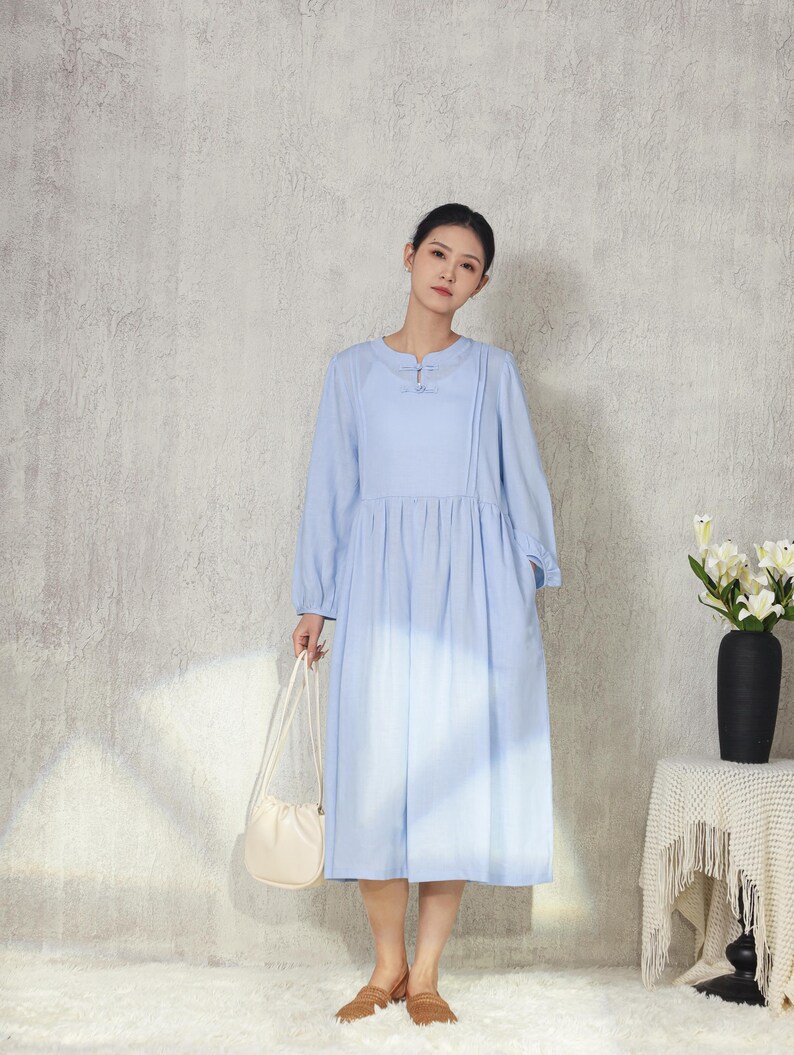 May include: A light blue dress with long sleeves and a round neckline. The dress has a gathered waist and falls to mid-calf. A small, off-white purse is held in the hand. The background is a light gray wall and a white rug.