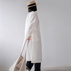 May include: A woman wearing a white linen dress with a straw hat and a beige tote bag.