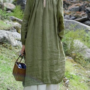 May include: A loose-fitting, long-sleeved, olive green linen dress with a round neckline and a small keyhole opening at the neckline. The dress has a gathered detail at the neckline and a gathered hemline. The model is wearing the dress with a white skirt and is holding a woven basket.