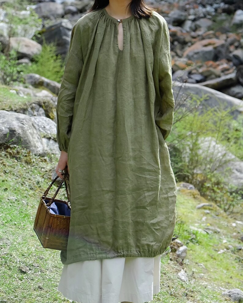 May include: A loose-fitting, long-sleeved, olive green linen dress with a round neckline and a small keyhole opening at the neckline. The dress has a gathered detail at the neckline and a gathered hemline. The model is wearing the dress with a white skirt and is holding a woven basket.